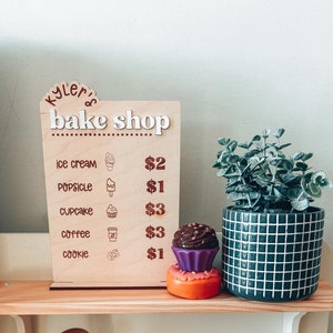 May include: A wooden sign with the words "kyler's bake shop" written on it. The sign lists the prices for ice cream, popsicle, cupcake, coffee, and cookie. The sign is sitting on a wooden shelf with a gold faucet and a potted plant.