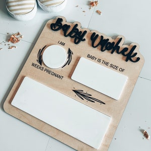 May include: A wooden baby tracker with white spaces to fill in the number of weeks pregnant and the size of the baby. The text "Baby Week" is written in black cursive lettering.