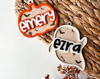 May include: Two Halloween-themed wooden ornaments. One is a pumpkin shape with the name "emery" in white, surrounded by bats and a skull. The other is a ghost shape with the name "ezra" in black, with bats and a bone.