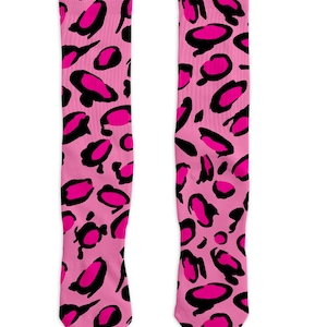 May include: A pair of knee-high socks with a pink leopard print design. The socks feature large, irregular black spots with bright pink centers against a solid pink background. The socks are displayed on a white background.