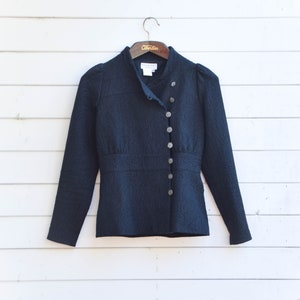 Könnte beinhalten: Marineblaue Jacke mit strukturiertem Muster, Stehkragen und langen Ärmeln. Die Jacke hat dekorative Knöpfe vorne und hängt an einem Holzbügel.