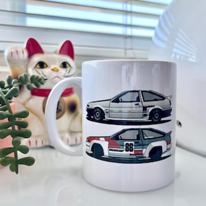 May include: White ceramic mug with a graphic of two white Toyota AE86 cars. The top car is a stock version and the bottom car is a race car with the number 86 on the side.