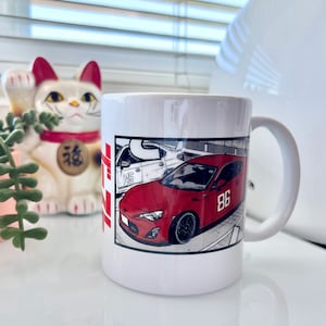 May include: White ceramic mug with a red and black graphic of a red sports car with the number 86 on the side. The car is in a parking lot with other cars in the background.