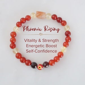 May include: A red and yellow beaded bracelet with a charm featuring an evil eye design. The bracelet is labeled "Phoenix Rising" and "Vitality & Strength, Energetic Boost, Self-Confidence".