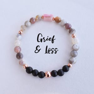 May include: A bracelet featuring black and grey agate beads, rose gold beads, and a pink stone charm. The bracelet is inscribed with the words "Grief & Loss".