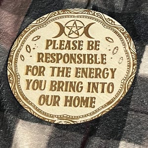 May include: A round wooden sign with a pentagram and crescent moon design. The sign reads "Please be responsible for the energy you bring into our home."
