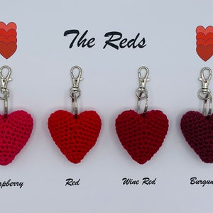 May include: Four crocheted heart-shaped keychains in different shades of red: raspberry, red, wine red, and burgundy. The keychains are labeled with their color names and the text "The Reds" is displayed at the top of the image.