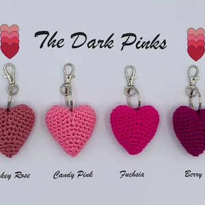 May include: Four crocheted heart-shaped keychains in different shades of pink, including smokey rose, candy pink, fuchsia, and berry. The keychains are labeled with their respective colors.