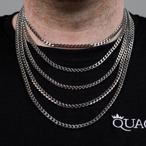 Könnte beinhalten: Eine silberne Kettenkette mit mehreren Lagen. Die Kette wird von einer Person getragen, die ein schwarzes Shirt mit dem Wort "QUAGEN" auf der Vorderseite trägt.