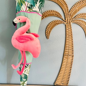 May include: A green and white cone-shaped school bag with a pink flamingo plush toy attached. The bag has a tropical leaf pattern and a pink and white trim. The bag is hanging on a wall with a wicker palm tree decoration.