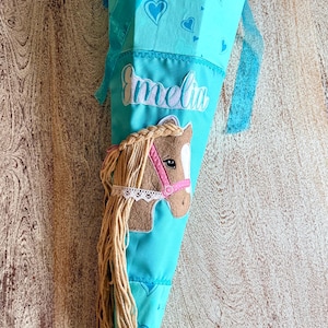 Könnte beinhalten: Eine türkisfarbene Schultüte mit einem Pferde-Applikation und dem Namen "Amelia" aufgestickt. Die Tüte ist mit Herzmustern und einem blauen Tüllaufsatz verziert. Das Pferd hat ein rosa Halfter und eine lange, fließende Mähne.