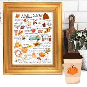 May include: A framed print with a gold border displays a fall bucket list with illustrations of autumn activities. The list includes items like drinking apple cider, visiting a pumpkin patch, and roasting pumpkin seeds. A coffee cup with a pumpkin graphic is in front.