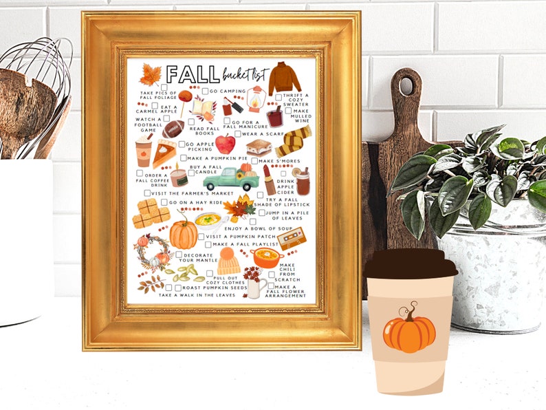 May include: A framed print with a gold border displays a fall bucket list with illustrations of autumn activities. The list includes items like drinking apple cider, visiting a pumpkin patch, and roasting pumpkin seeds. A coffee cup with a pumpkin graphic is in front.