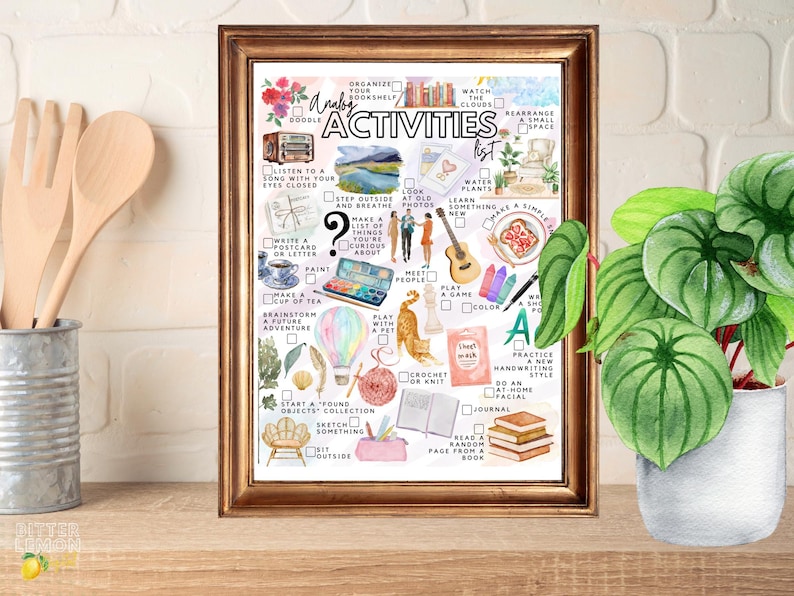 May include: A framed print with a gold-colored frame features a watercolor-style illustration of an "Analog Activities List." The list includes activities like "Listen to a song," "Paint," and "Read a random page from a book." A potted plant sits to the right.