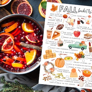 May include: An overhead shot shows a pot of mulled wine with orange slices and cranberries, next to a "Fall Bucket List" with autumn activities. The list includes apple picking, pumpkin pie baking, and enjoying fall foliage.