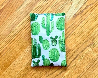 May include: A white fabric pouch with a green cactus print. The pouch is filled with a soft material, likely for use as a heating pad or eye mask.