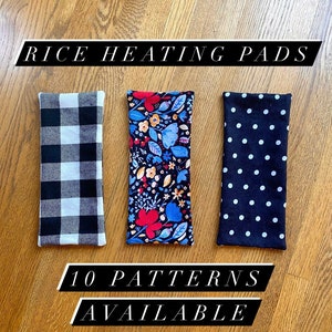 May include: Three rectangular rice heating pads with different patterns. The pads are black with white checkered, floral, and polka dot patterns. The text "RICE HEATING PADS" is at the top of the image and the text "10 PATTERNS AVAILABLE" is at the bottom of the image.