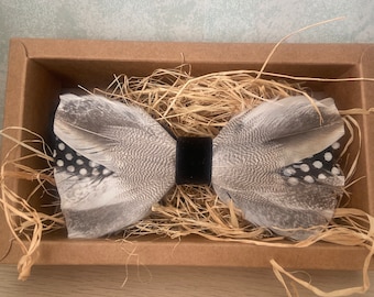 Könnte beinhalten: Handgefertigte Fliege aus grau-weißen Federn mit schwarzen Punkten, in einer braunen Pappschachtel mit Stroh. Die Fliege hat eine schwarze Samtmitte.