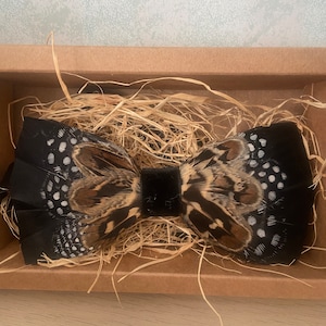 Puede incluir: Una pajarita de plumas negras con detalles moteados marrones y blancos, presentada en una caja de cartón marrón con paja. La pajarita tiene un centro de terciopelo negro, ideal para eventos formales.