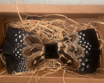 Könnte beinhalten: Eine schwarze Feder-Fliege mit braunen und weißen gesprenkelten Akzenten, präsentiert in einer braunen Pappschachtel mit Stroh. Die Fliege hat eine schwarze Samtmitte und eignet sich für formelle Anlässe.