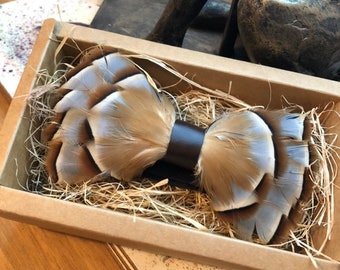 May include: A brown and white feather bow tie with a brown leather strap. The bow tie is in a brown box with straw filling.