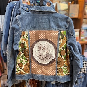 Pode incluir: Jaqueta jeans com painel marrom de bolinhas e ilustração circular de cogumelos. Detalhes em tecido floral marrom, verde e amarelo emolduram o desenho de cogumelos. Outras jaquetas jeans são visíveis ao fundo.