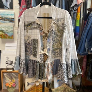 Puede incluir: Chaqueta patchwork con un patrón de cuadrícula blanca, adornada con parches de tela floral y de mariposas. La chaqueta tiene un dobladillo con volantes y mangas largas. La chaqueta está colgada en una percha negra.