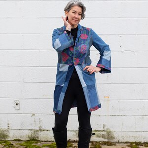 May include: A patchwork denim coat with floral and striped fabric accents. The coat features a long, tailored design with a button closure and bell sleeves. The model is wearing black leggings and black boots.