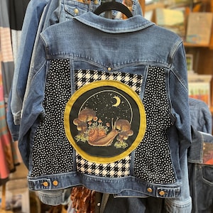 May include: Blue denim jacket with a unique design on the back. The design features a circular patch with a snail, mushrooms, and a crescent moon. The jacket has black and white polka dot and houndstooth patterns.