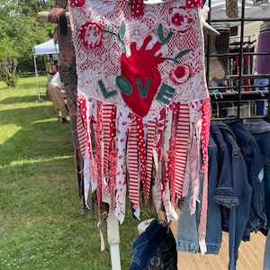 May include: A handmade wall hanging with a red heart and the word "LOVE" appliqued on a white lace background. Red and white fabric strips hang from the bottom. The top has red polka dot ribbon and is suspended from a wooden rod.