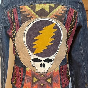 May include: Dark blue denim jacket with a colourful, patterned back panel. The back features a large patch with a skull and lightning bolt design. The jacket has a classic collar and button closures. The back panel has a geometric design in shades of red, orange, and brown.