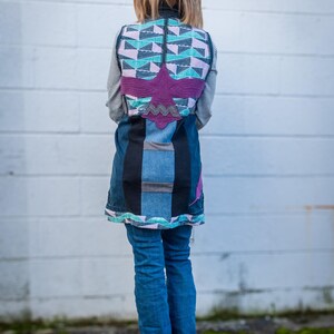 Könnte beinhalten: Eine Patchwork-Jeansweste mit einem geometrischen Design in Blau-, Türkis- und Lavendeltönen. Die Weste hat eine große Applikation eines lila Vogels und wird über einem gestreiften Langarmshirt und einer blauen Jeans getragen.