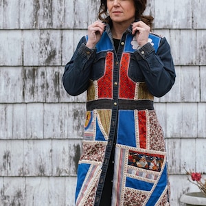 Puede incluir: Un abrigo largo de mezclilla patchwork con una variedad de patrones y colores de tela, incluyendo rojo, azul y marrón. El abrigo tiene una parte delantera abotonada y se usa sobre una camiseta negra. El fondo es una pared de madera blanca desgastada.