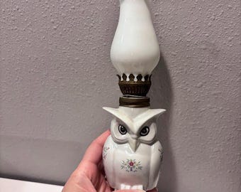 Puede incluir: Una lámpara de aceite de cerámica blanca con forma de búho y un patrón floral en el cuerpo. La lámpara cuenta con un quemador de metal color latón y una chimenea de vidrio blanco translúcido con un borde superior ondulado y vertical.