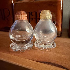 May include: Two clear glass shakers with rounded bodies and a base featuring a ring of small glass spheres. One shaker has a textured copper-colored lid, while the other has a translucent yellow lid. Both containers have a smooth, bulbous shape.