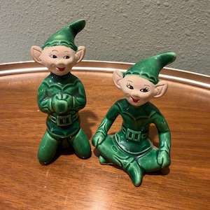 May include: Two ceramic elf figurines with pointed green hats and matching green outfits. One elf is kneeling with hands clasped, while the other sits cross-legged. Both have pale faces, pointed ears, and red painted lips.
