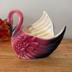 May include: A ceramic swan-shaped planter or dish featuring a glossy finish with a gradient of deep magenta and pink tones. The swan has a curved neck, textured wing details, and a cream-colored interior. The beak and wing tips are accented in black.