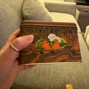 May include: A rectangular metal tin featuring a dark brown wood grain pattern with a painted design of orange strawberries, green leaves, and white flowers. The lid is a solid dark brown color. A wooden ruler is visible at the bottom of the frame.