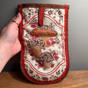 May include: A rectangular fabric oven mitt featuring a cream-colored background with a repeating pattern of brown and orange mushrooms, small brown flowers, and red leaves. The mitt is bordered by red and brown fabric trim and has a hanging loop.
