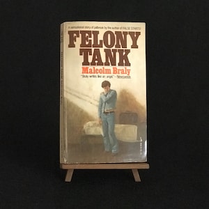 May include: A paperback book titled "Felony Tank" by Malcolm Braly. The cover features a painting of a young man standing in a room with a suitcase. The book is on a wooden easel.