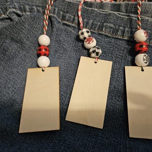 May include: Three wooden tags with decorative beads and twine. The beads feature white, red, and black patterns, including buffalo check and snowflakes. The tags are rectangular with a hole at the top for the twine.