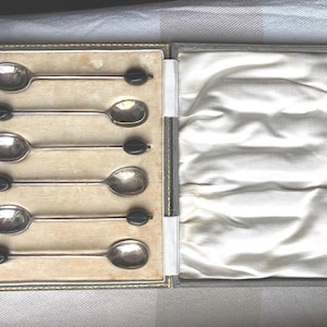 Könnte beinhalten: Ein Set aus sechs silbernen Teelöffeln mit schwarzen Griffen in einer beigefarbener Samtbox. Die Box ist geschlossen und die Löffel sind in Reihen angeordnet.