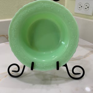 Puede incluir: Un cuenco de vidrio verde claro, de estilo vintage, con borde festoneado y diseño floral en relieve, exhibido en un soporte de metal negro. El cuenco es redondo y tiene un acabado liso y brillante. El soporte tiene adornos decorativos.