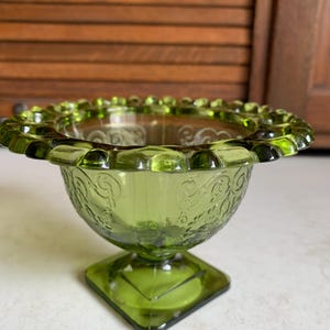 May include: A green glass bowl with a scalloped rim and a square base. The bowl has a delicate floral design.
