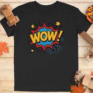 以下が含まれることがあります： 黄色、赤、青のコミック風「WOW！」グラフィックが特徴の黒いTシャツ。デザインには星と吹き出しが含まれています。シャツは、秋をテーマにした装飾が施された木製の表面に表示されています。