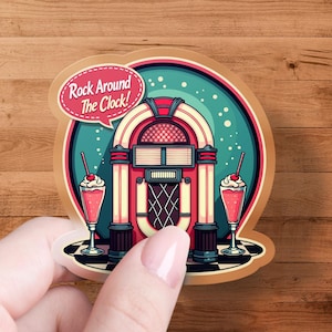 May include: A retro-style sticker featuring a jukebox design with the text "Rock Around The Clock!". The sticker also includes illustrations of two pink milkshakes with cherries. The background is teal and the border is brown.