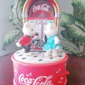 May include: A Coca-Cola themed musical carousel with two white teddy bears. The bears are dressed in red and blue outfits and are facing each other. The carousel is red and white with the Coca-Cola logo on the base.
