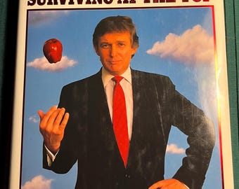 May include: A book cover with the title "TRUMP SURVIVING AT THE TOP" in red letters. The cover features a photo of Donald J. Trump in a black suit and red tie, holding an apple in his hand. The background is a blue sky with white clouds.