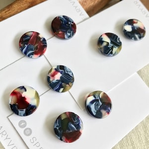 May include: Seven circular buttons featuring a marbled pattern in shades of deep red, navy blue, white, and translucent grey. Each button has a smooth, glossy finish and two small holes for attachment, displayed on white branded display cards.