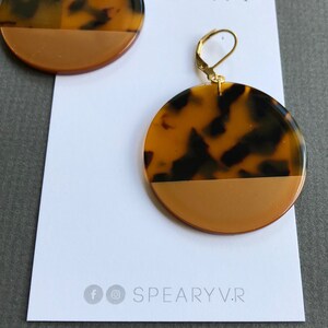 May include: A pair of round earrings featuring a tortoiseshell pattern on the top half and a solid mustard yellow color on the bottom half. The earrings are attached to a white display card with the text SPEARYV.R and social media icons.